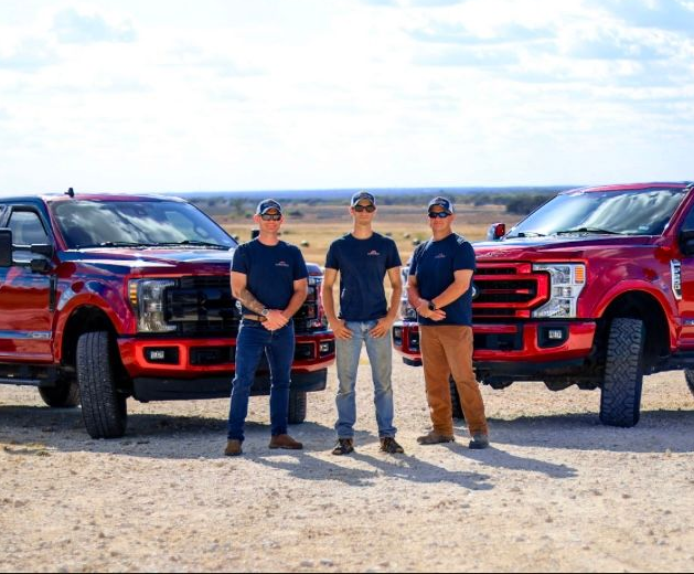 Three men stand between two red trucks on a dirt road, sunny sky in the background.