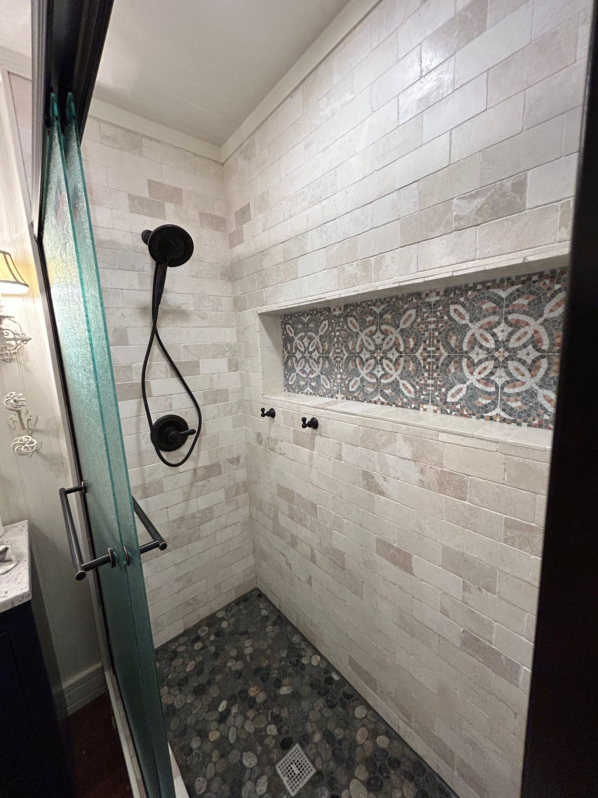 Shower with glass door, pebble floor, white tile walls, decorative niche, black showerhead.