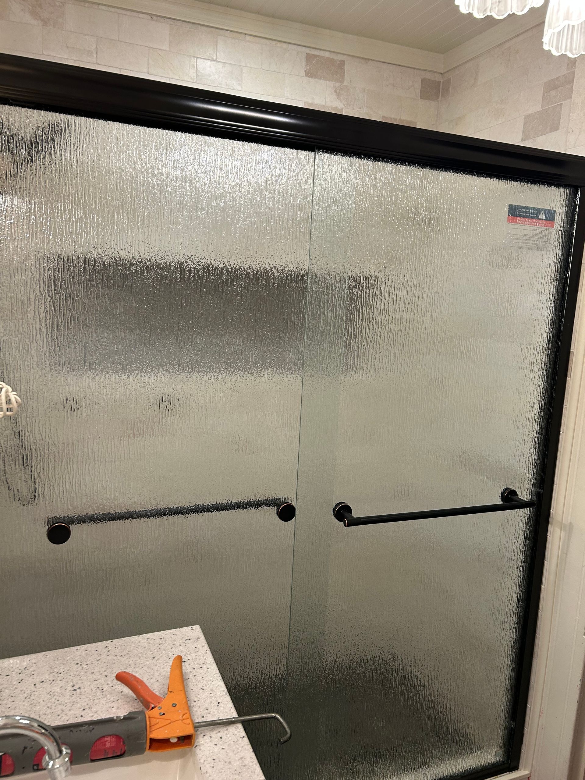 Shower with textured glass doors, black frame, and black handles. A caulk gun rests on the counter.