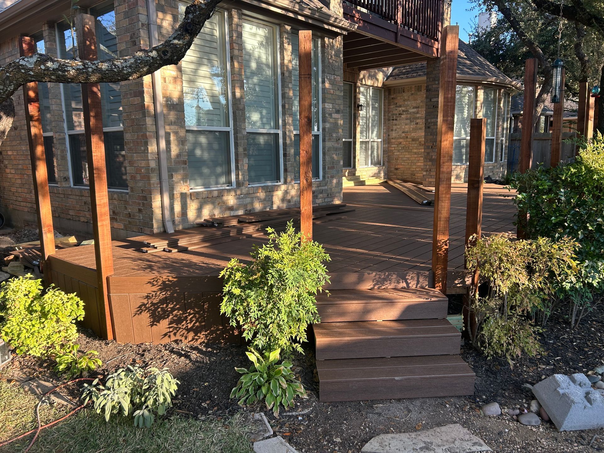 Brown wooden deck with steps leading to a house, surrounded by landscaping and trees.