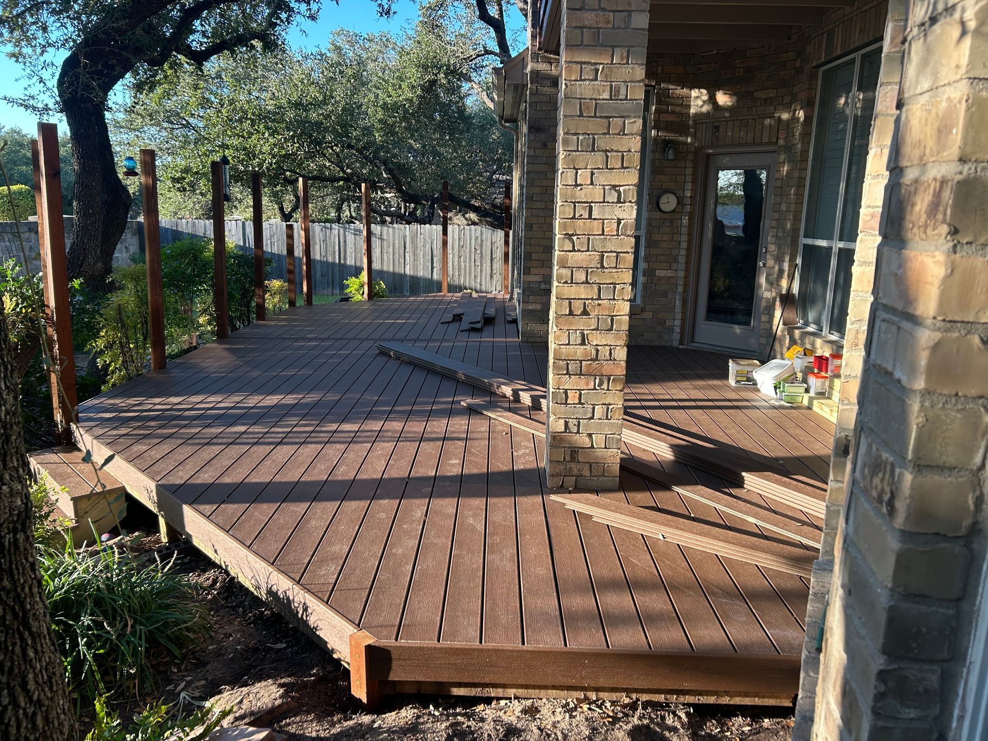 Deck under construction with brown composite decking. Brick columns, wooden fence, and trees in the background.