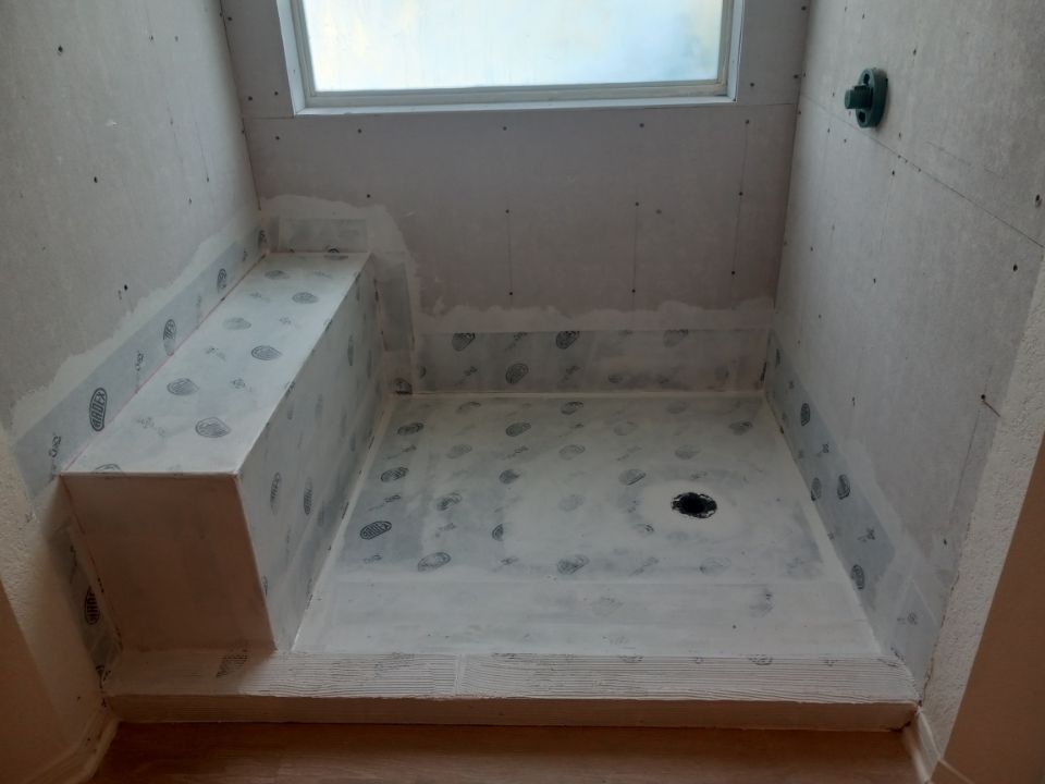 A shower stall under construction, with a bench, skylight, and drain. Gray and white surfaces.