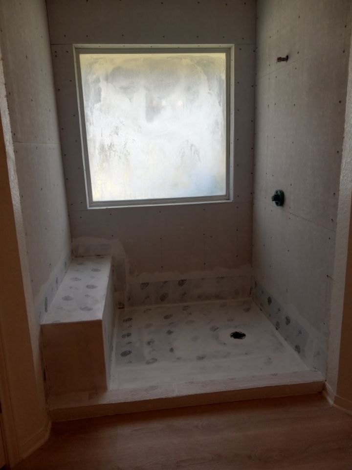 Shower stall under construction, with a bench, window, and drain. Drywall walls, light-colored flooring.
