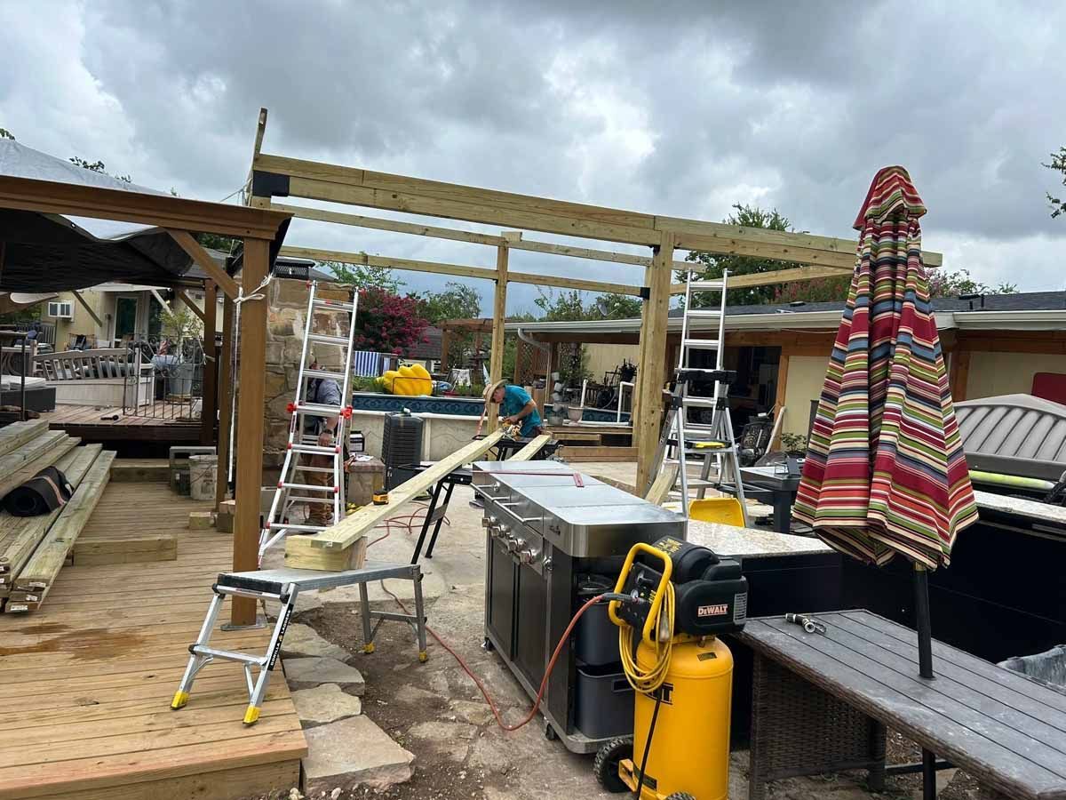 Construction of an outdoor wooden structure. People work on the frame with ladders, tools, and materials under a cloudy sky.