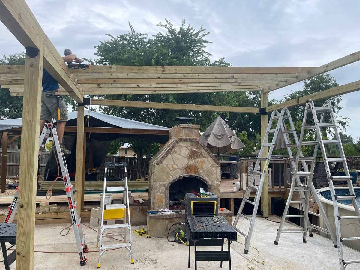 Construction of a wooden pergola in a backyard, with workers on ladders using tools near an outdoor fireplace.