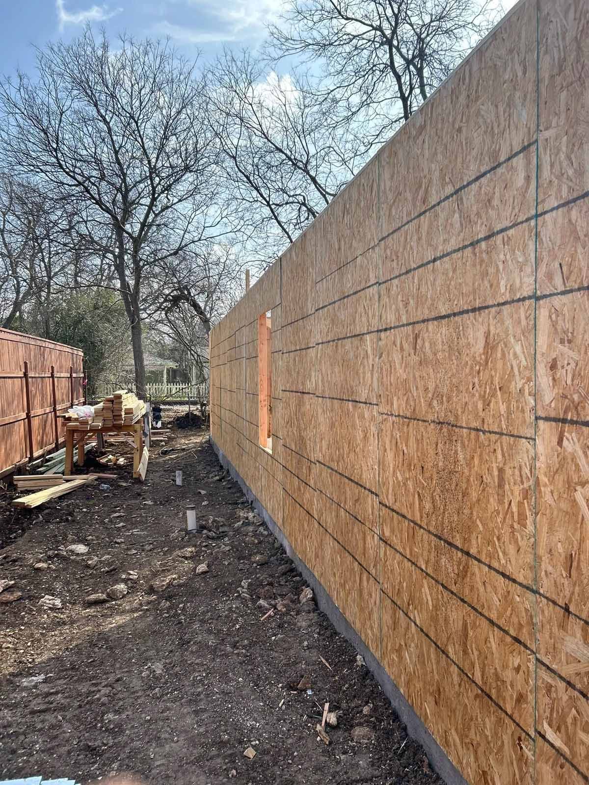 Construction site with wood-paneled wall and small window. Ground is dirt, trees in background, sunny day.