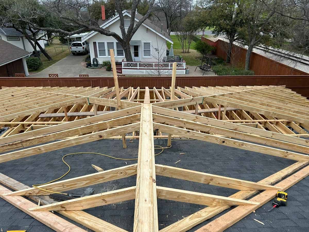 Wooden roof frame under construction with background of houses and sky.