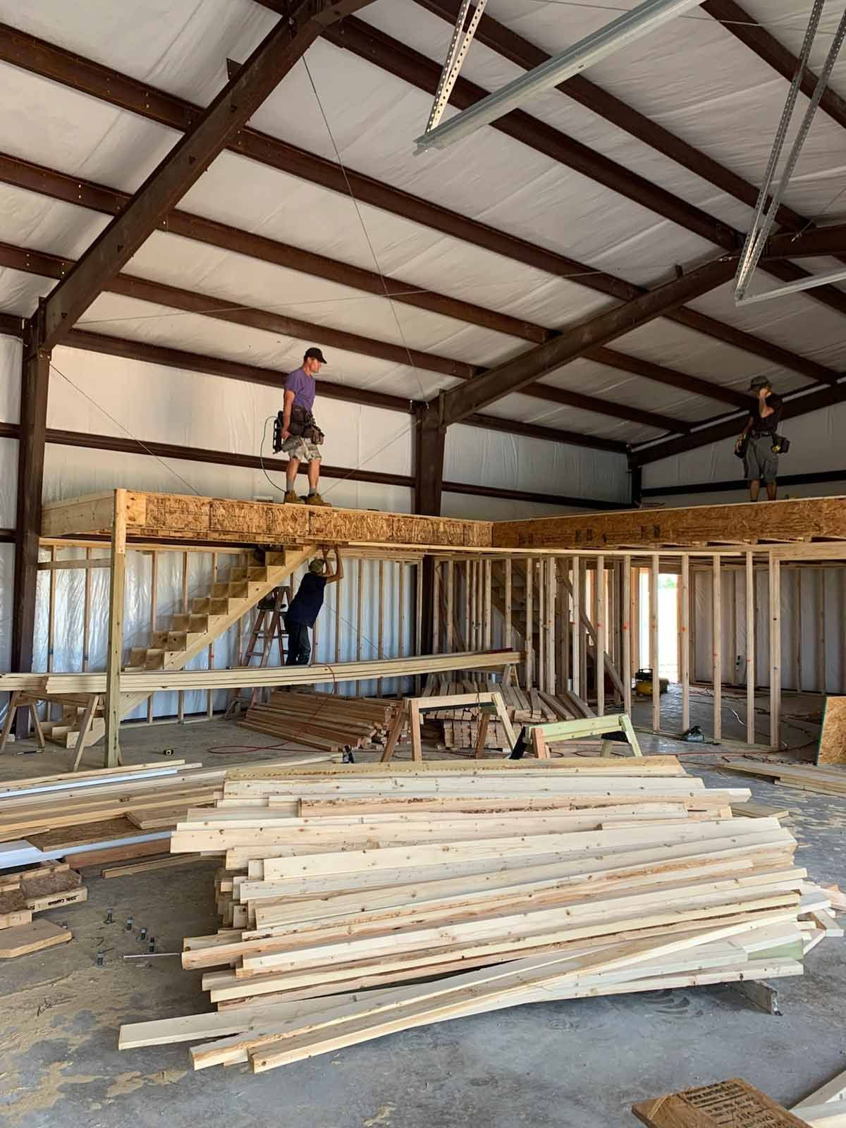 Construction of a mezzanine in a large metal building. Workers stand on framing, with lumber and stairs visible.