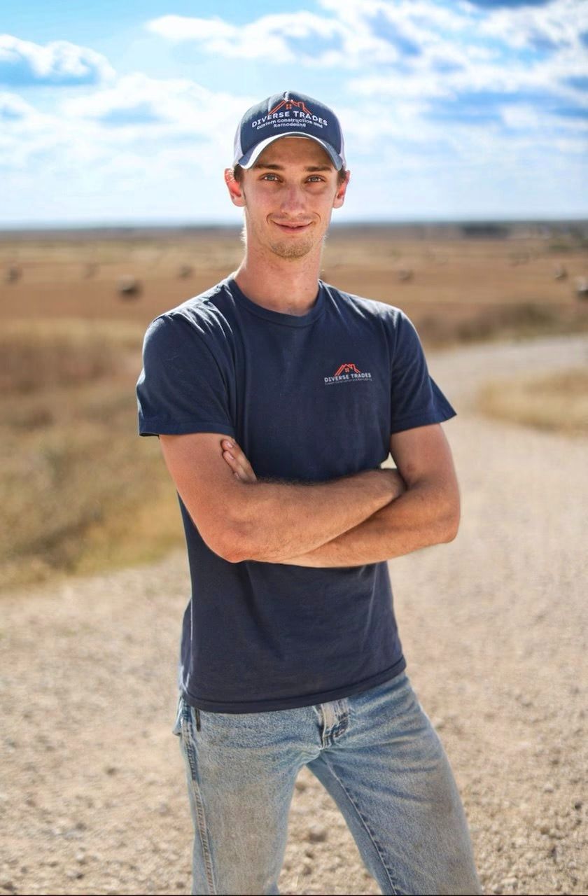 Man in blue shirt and hat, arms crossed, standing on dirt road in rural setting.