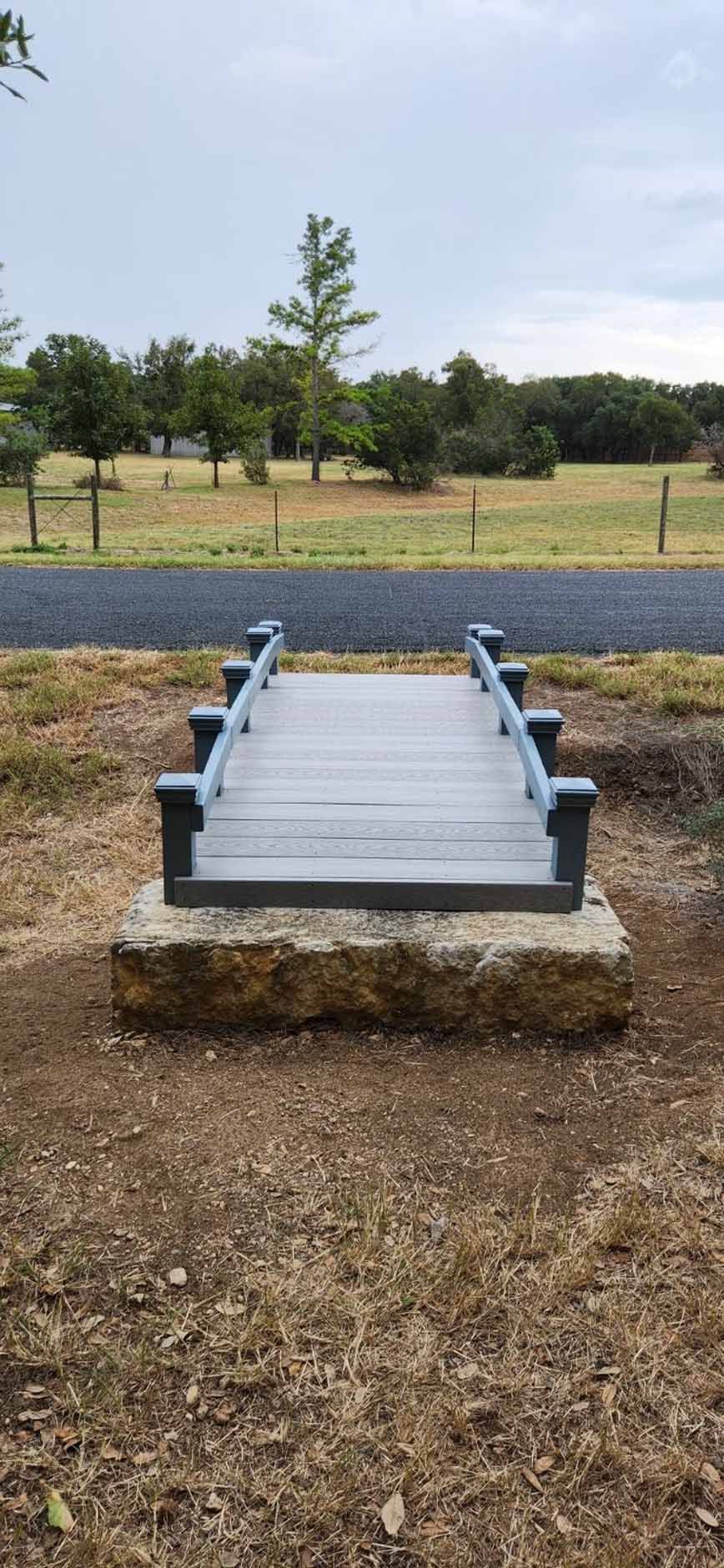 Miniature wooden bridge on a stone base, set in a grassy field with trees in the background.