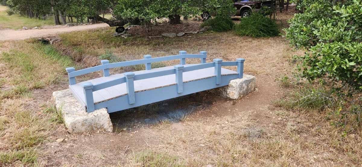 A small blue bridge over a dry creek bed in a grassy area, with trees in the background.