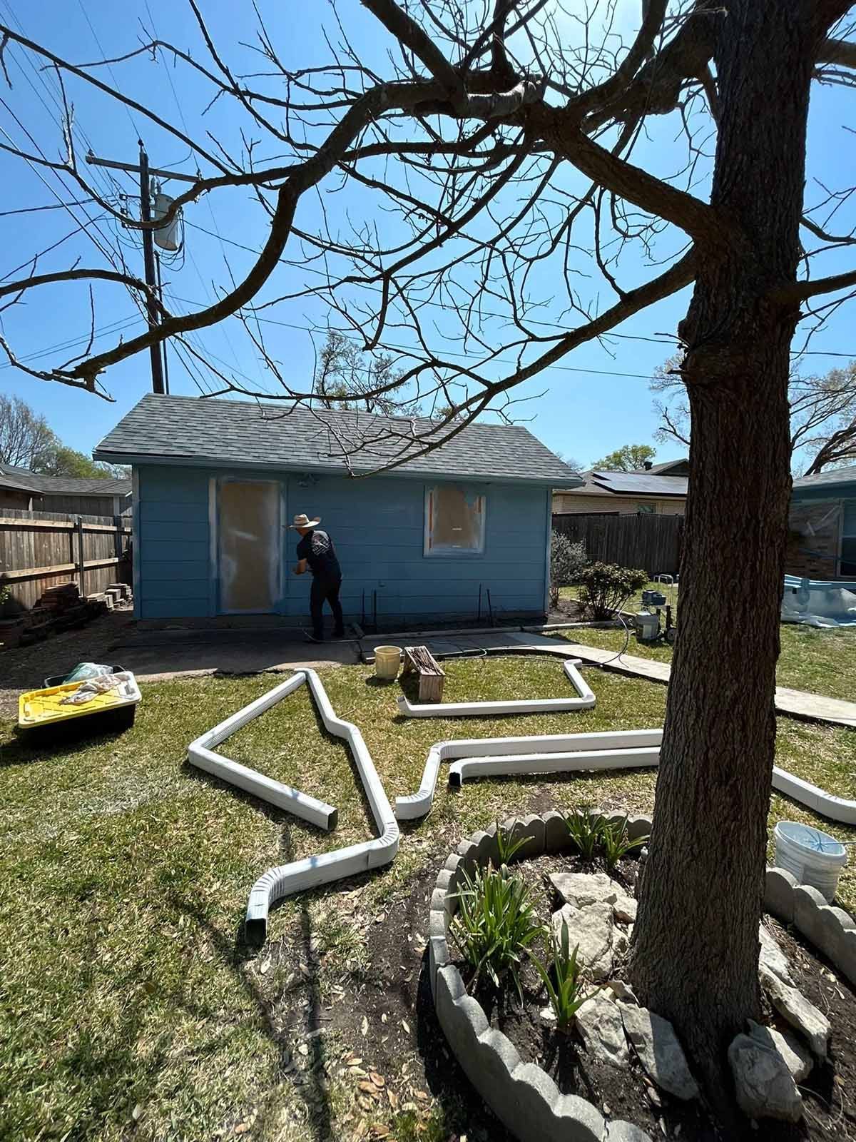 Backyard scene with a light blue building, a person working, and white gutter pieces on the grass.