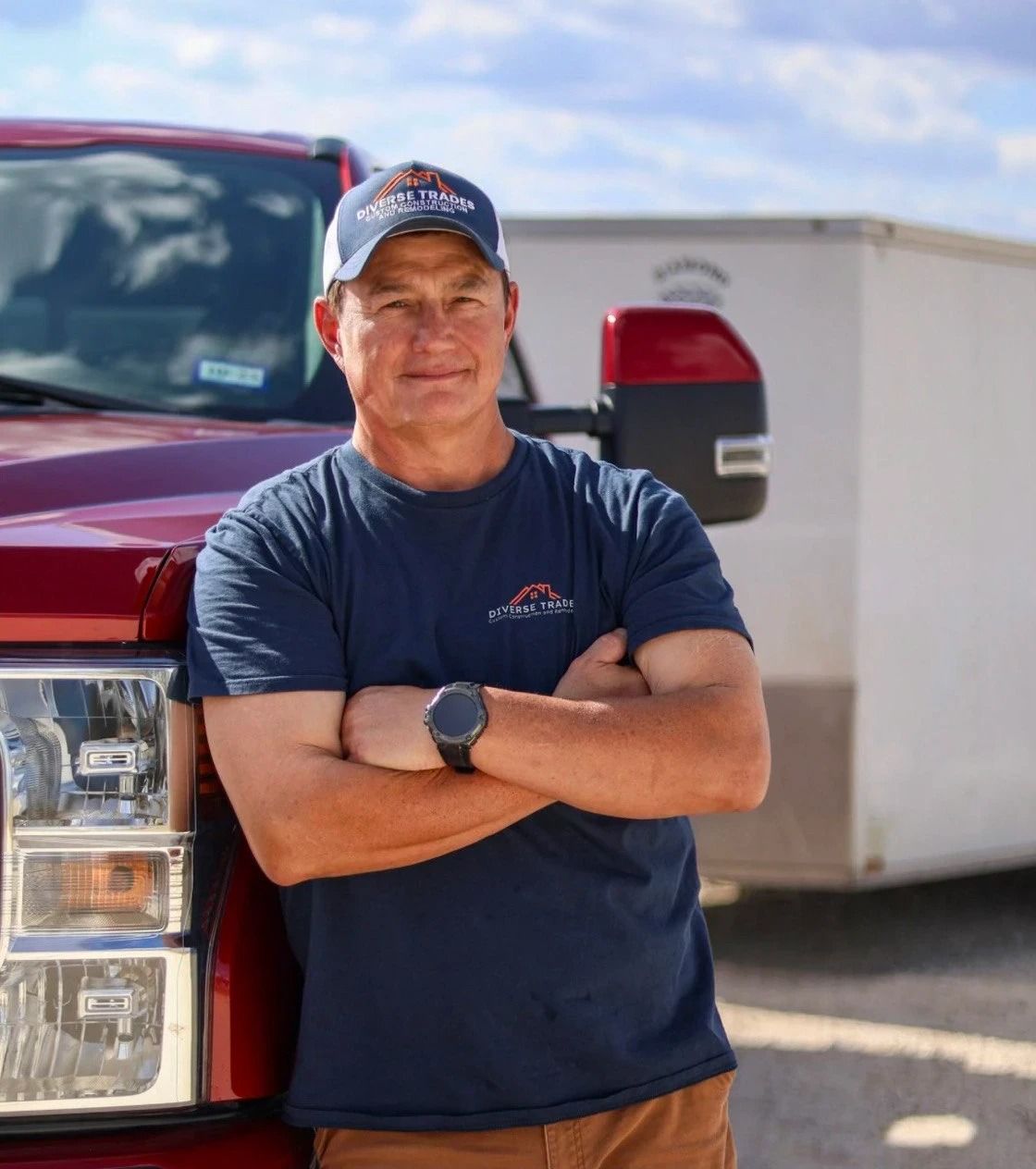 Man in a blue shirt and baseball cap, arms crossed, leaning against a red truck, with a trailer in the background.