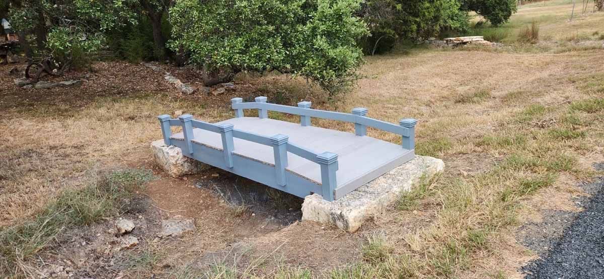 Small decorative blue wooden bridge over a shallow dry ditch, surrounded by dry grass and trees.