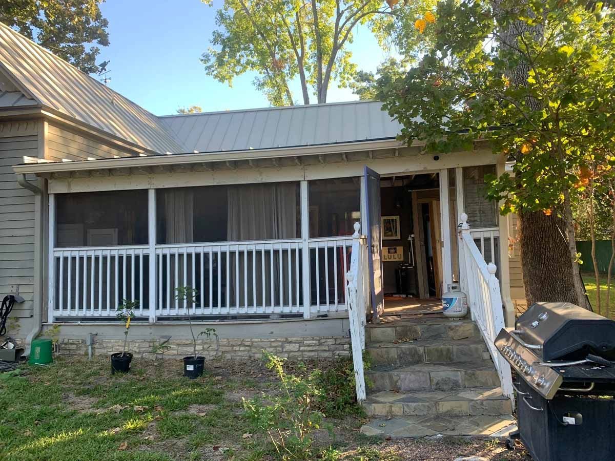 Screened porch with white railings and steps, leading to an open doorway. Metal roof and trees.