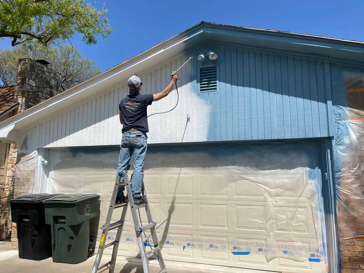 Man on ladder painting a garage door blue with a paint sprayer.