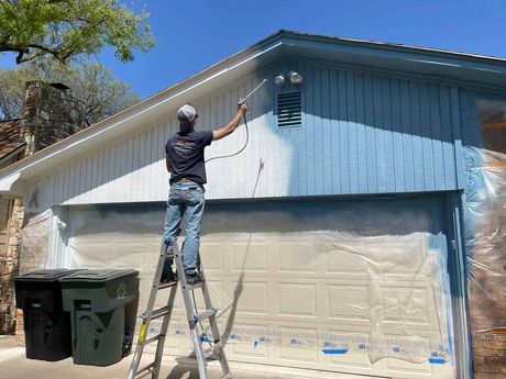 Man on ladder painting a garage door blue with a paint sprayer.