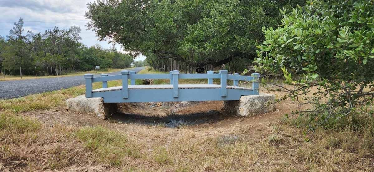 A blue wooden bridge over dry grass, trees in the background, gray asphalt road to the left.