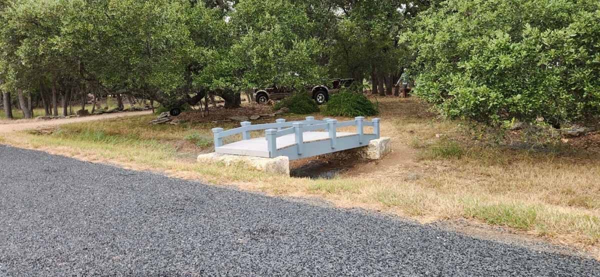 A small blue-grey bridge over dry grass, trees in the background, a gravel path in foreground.