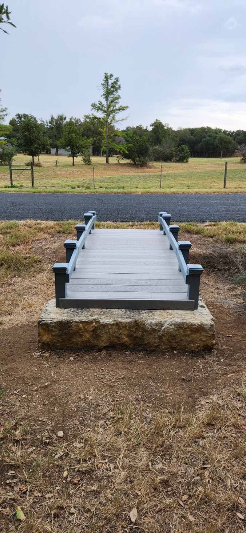 A small wooden bridge over a dirt area, with trees and a cloudy sky in the background.