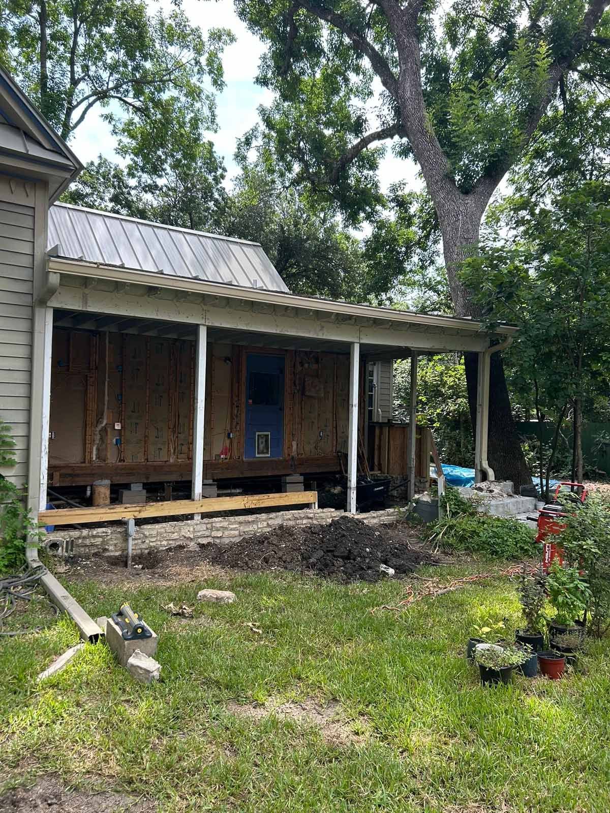 Exterior of a house with a porch under construction. Green grass, trees, and sky are visible.