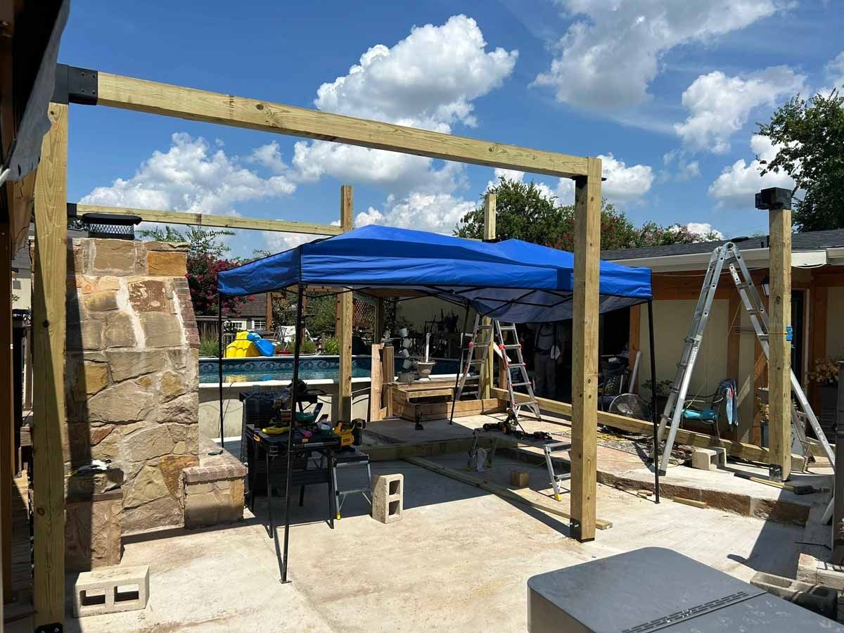 Construction of a wooden pergola frame over a patio area with a blue canopy, near a pool.
