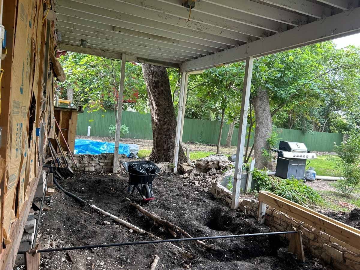 Construction site under a covered porch. Excavated dirt, wood framing, and a grill in the background.