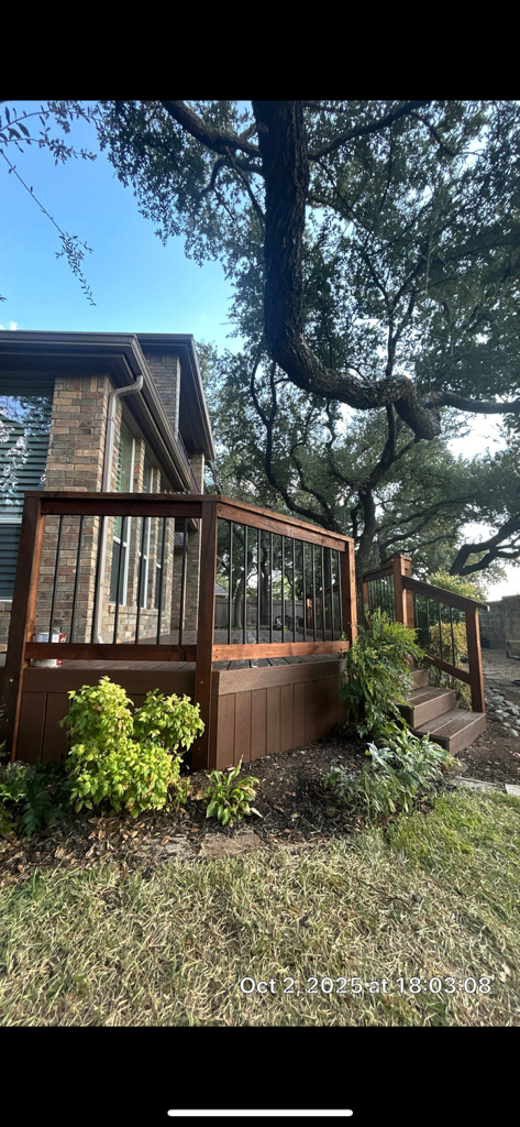 A deck with a brown railing next to a brick building, lush green foliage in the foreground.