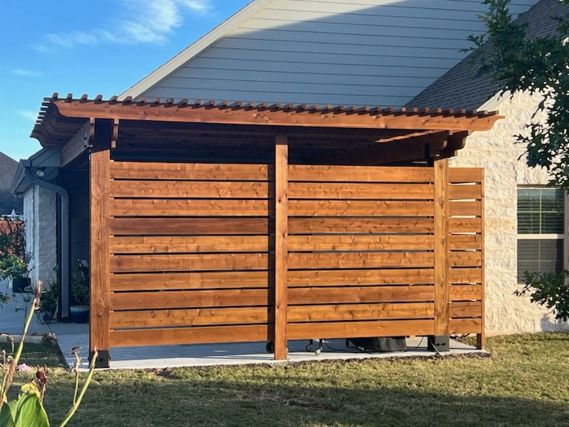 Wooden outdoor structure with a slatted privacy wall, and a corrugated roof, against a house.
