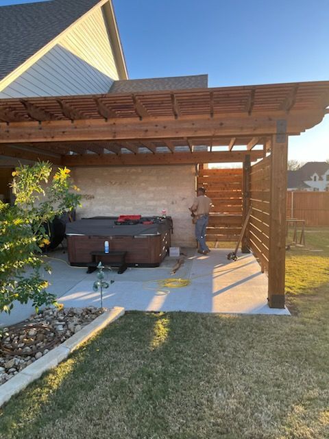 Wooden pergola over a hot tub on a concrete patio. A person is working in the shaded area.