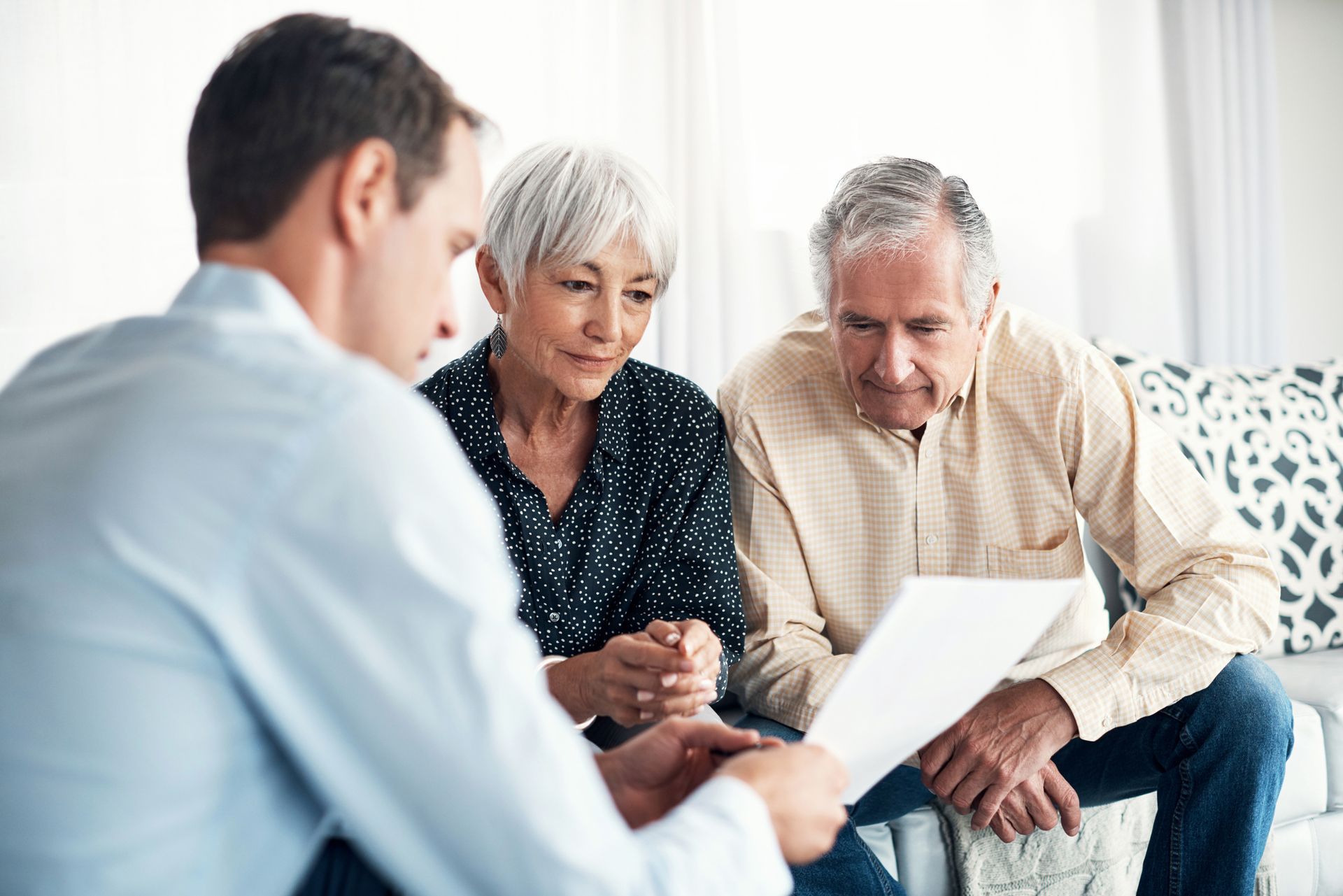 An elderly couple is sitting on a couch talking to a man while looking at a piece of paper.