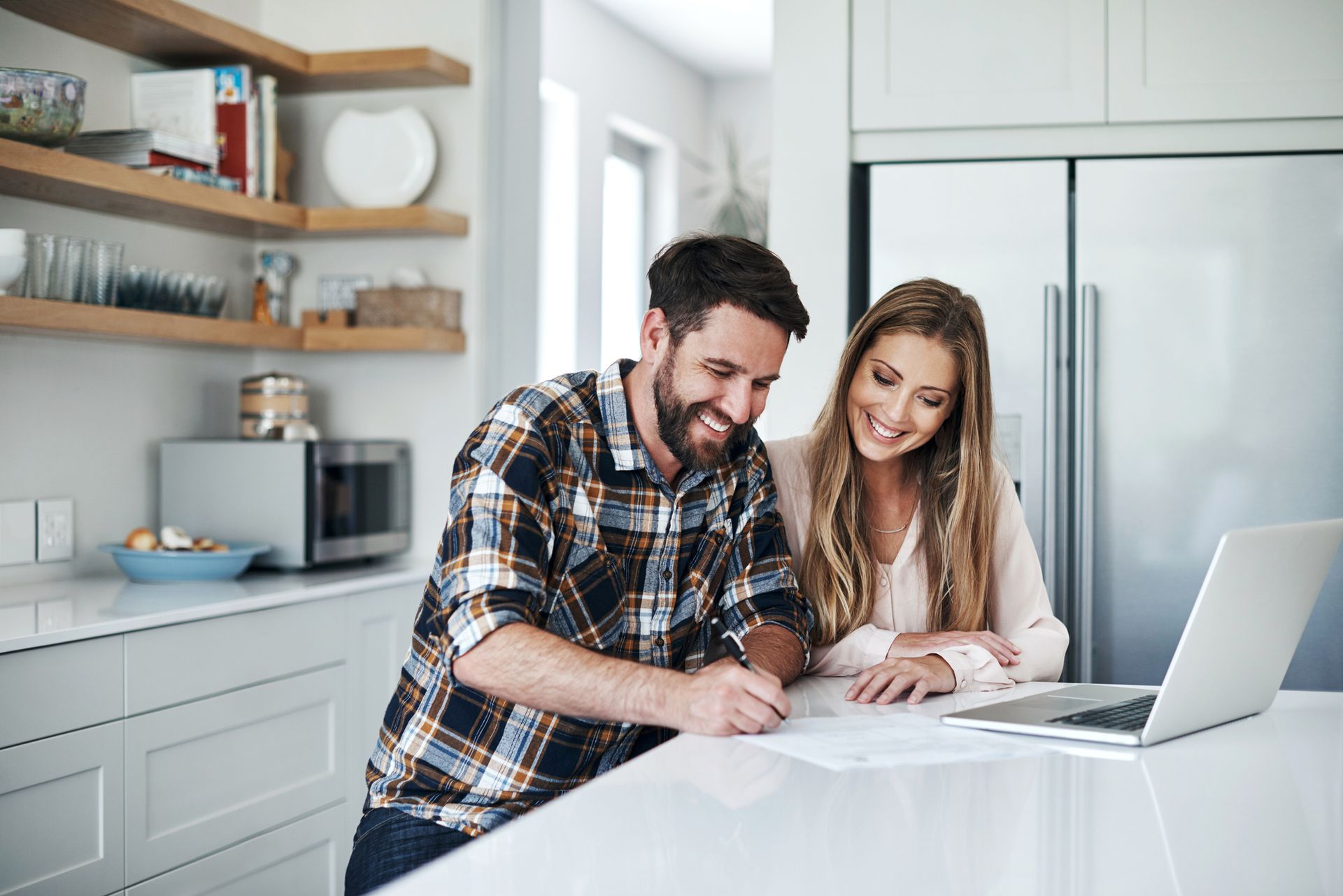 A man and a woman are sitting at a kitchen counter looking at a laptop.