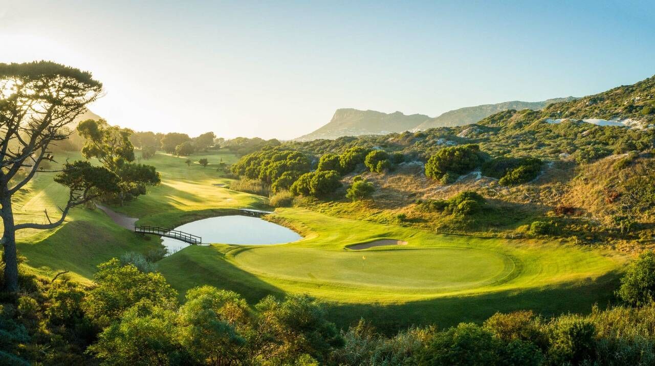 Trou de golf vert avec un étang au premier plan, des collines vallonnées et du soleil en arrière-plan.