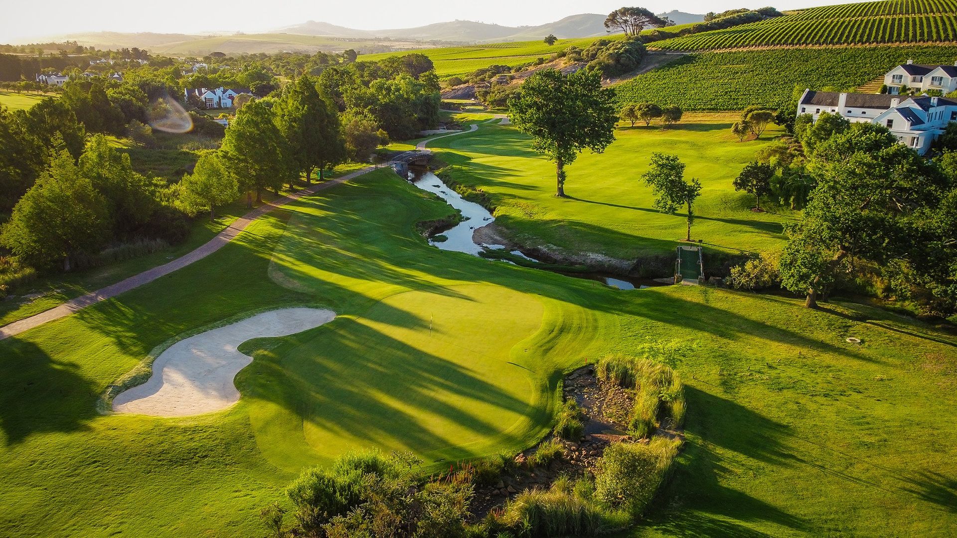 Terrain de golf verdoyant avec un bunker, un ruisseau et des maisons dans un paysage vallonné et ensoleillé.