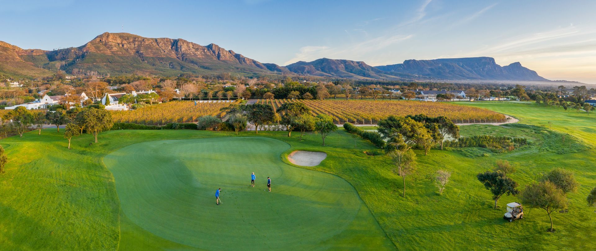 Des golfeurs sur un parcours verdoyant, des montagnes en arrière-plan, un ciel bleu, une lumière dorée.