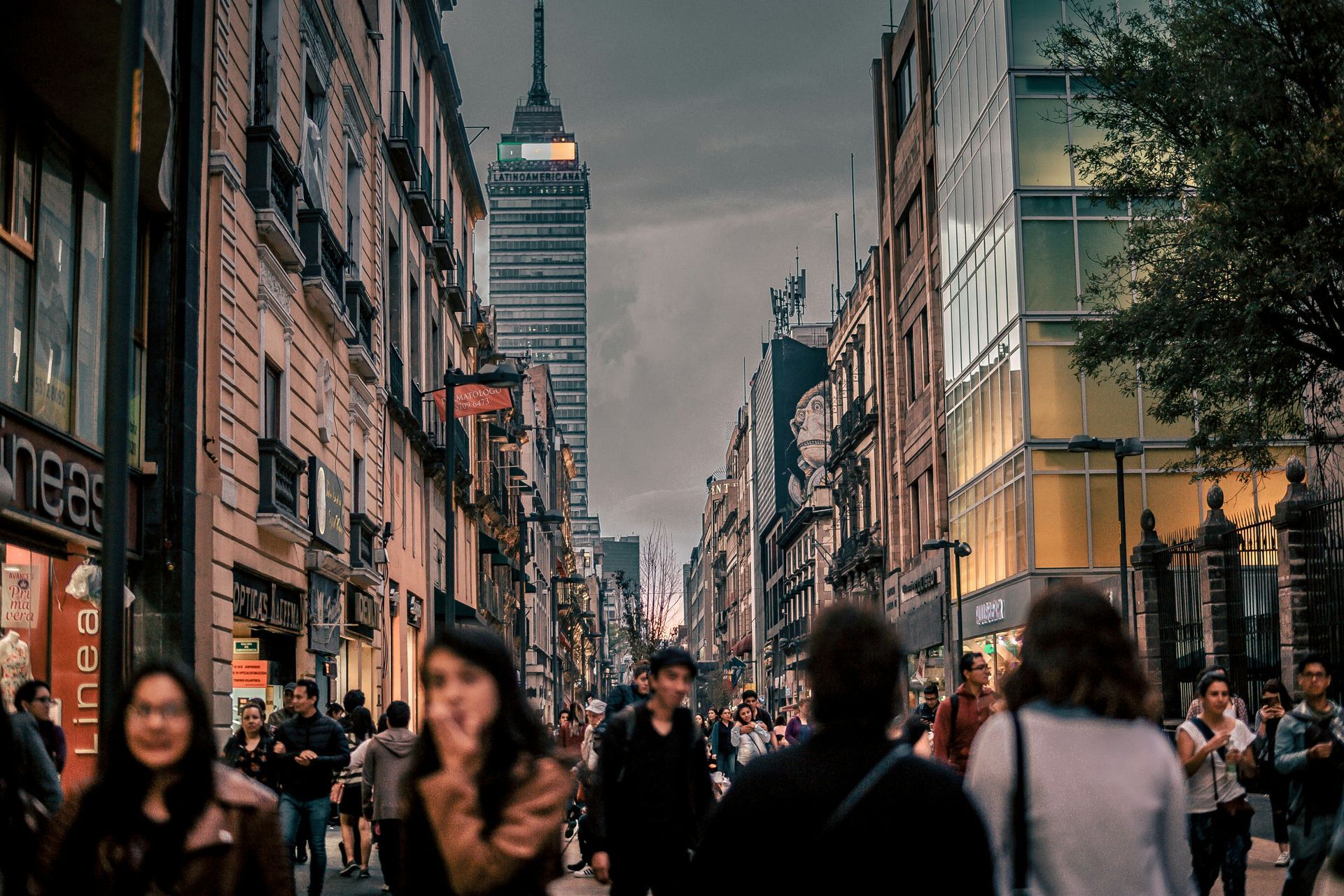 Scène de rue à Mexico, avec une foule de personnes marchant, une tour de grande hauteur visible au bout de la rue.