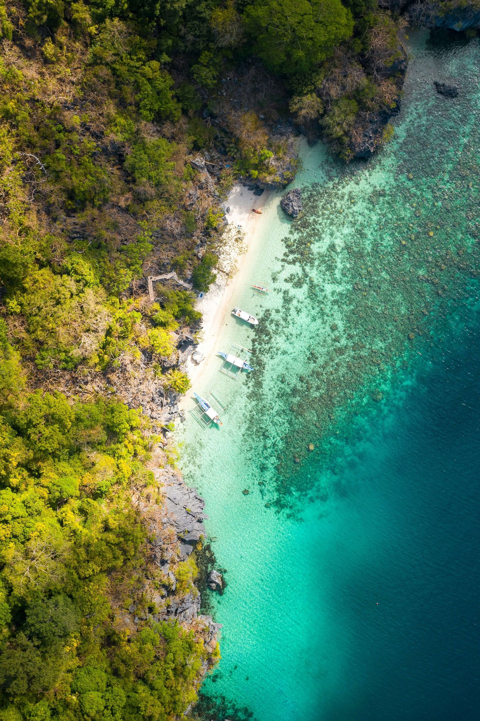 Vue aérienne d'une côte tropicale avec une eau turquoise, une plage de sable fin, des bateaux et une végétation luxuriante.