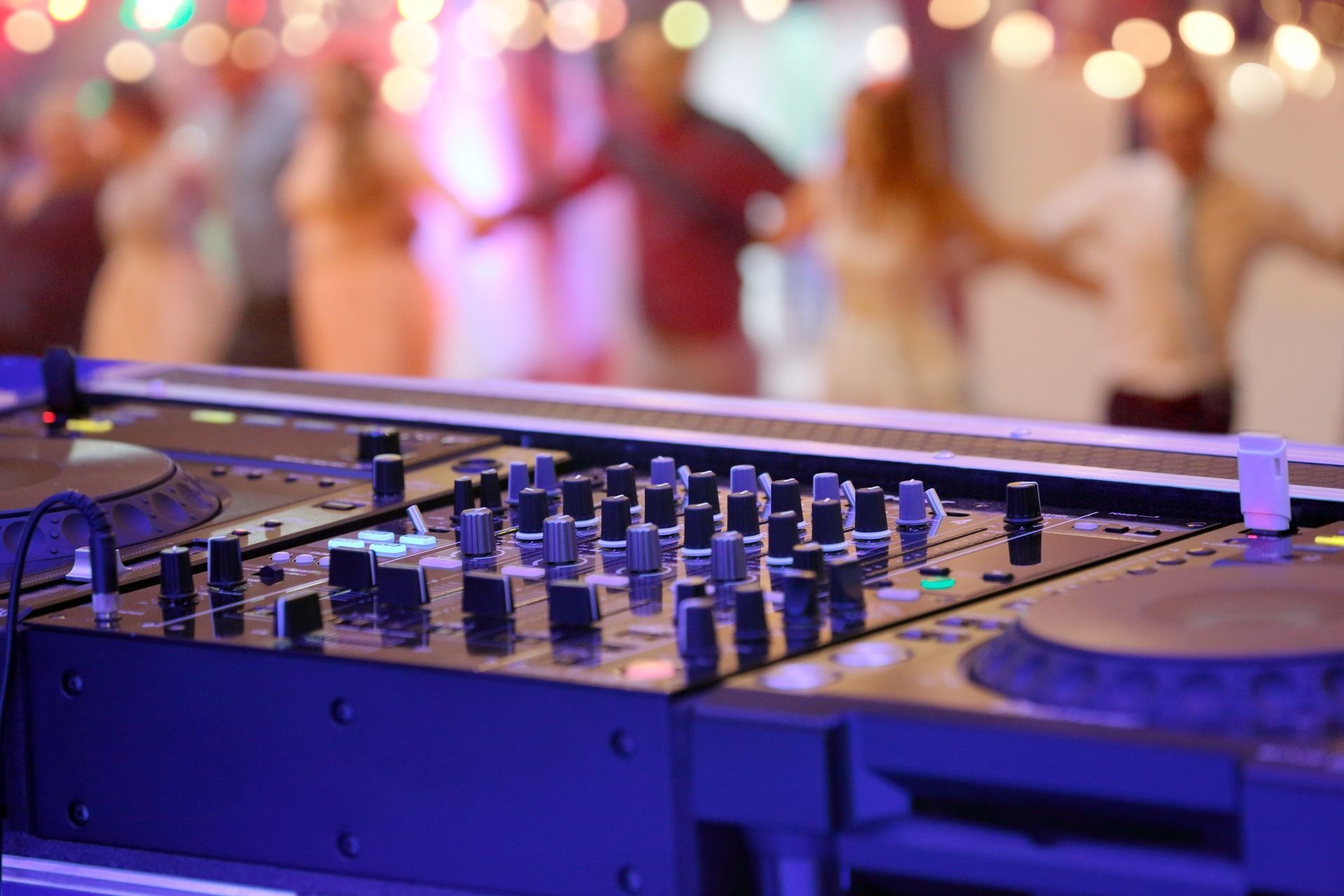 DJ mixer with dials and knobs in focus, blurred people dancing in the background with string lights.