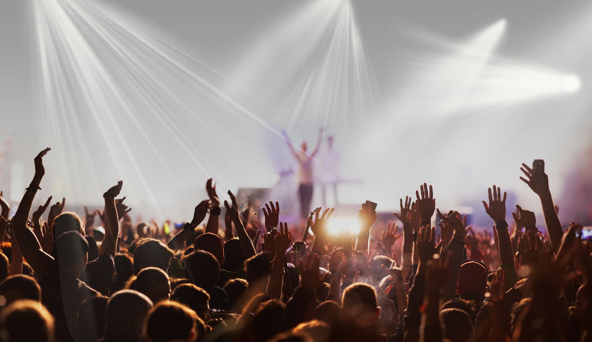 Concert crowd with raised arms, bright stage lights shining on a band in silhouette.