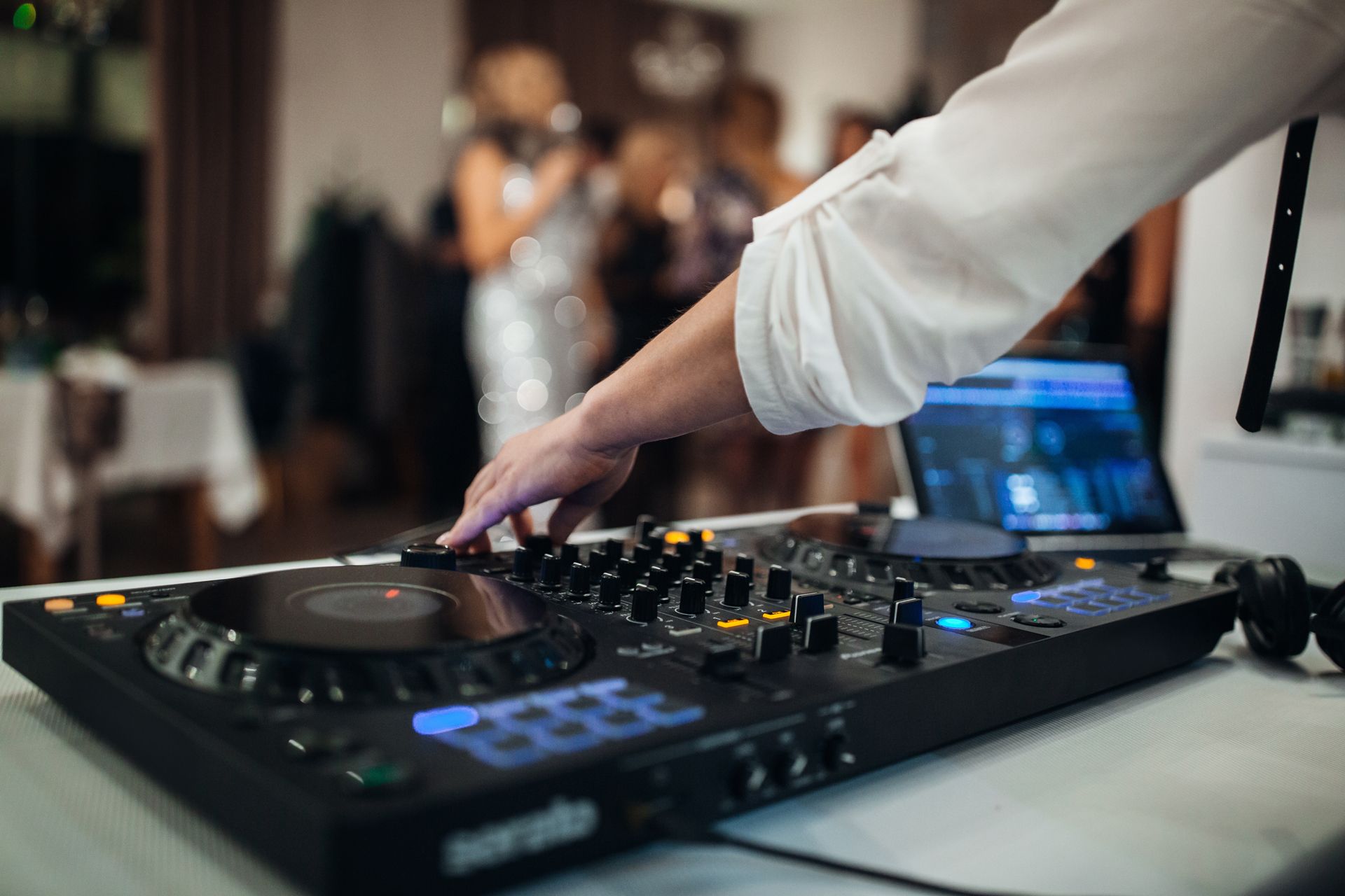 DJ at a party using a mixer, playing music, with blurred people in the background.
