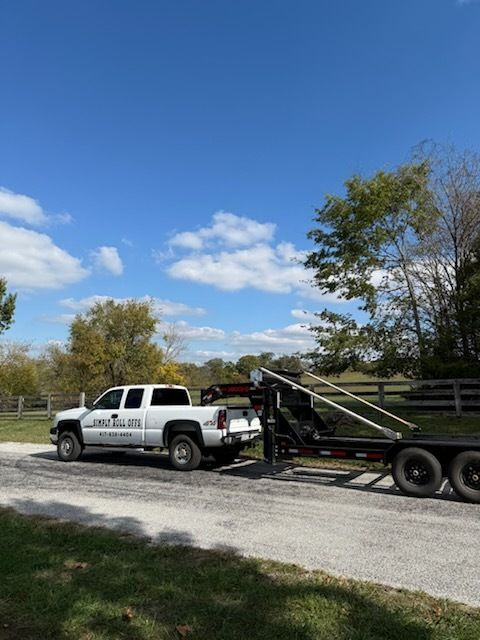 White pickup truck towing a trailer on a gravel road under a blue sky, trees in the background.