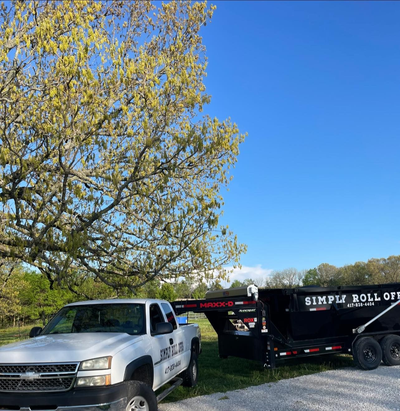 White pickup truck towing a black Simple Roll Off Services trailer under a tree, against a blue sky.