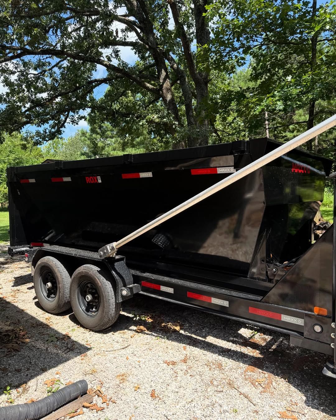 Black dump trailer parked outdoors on gravel with trees in the background.