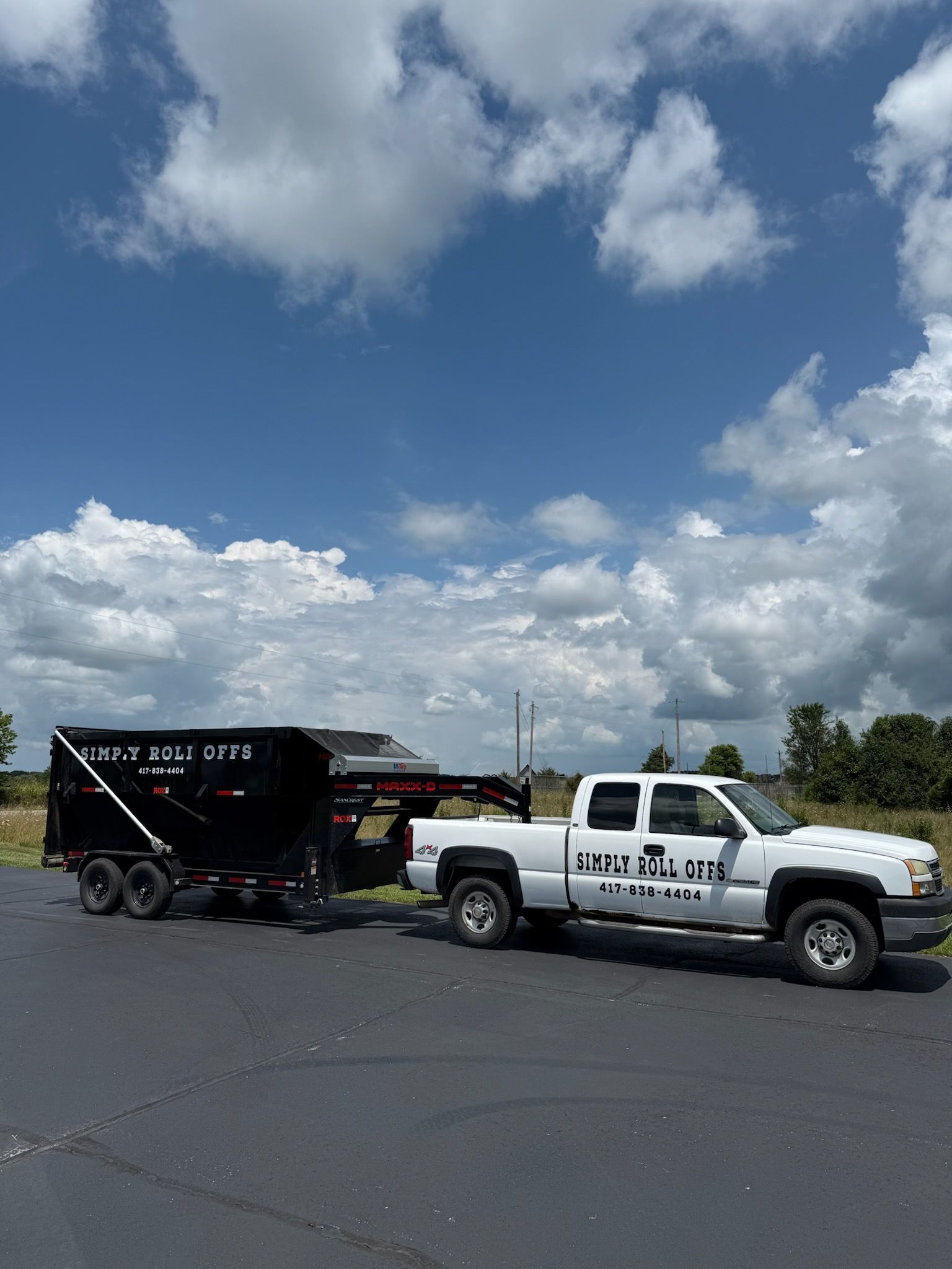 White truck towing a black dumpster trailer under a partly cloudy blue sky.