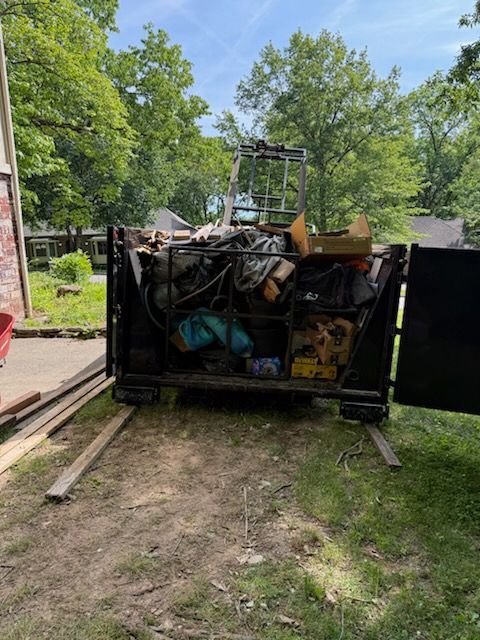 Overflowing dumpster in a grassy yard with construction debris, trees, and a house in the background