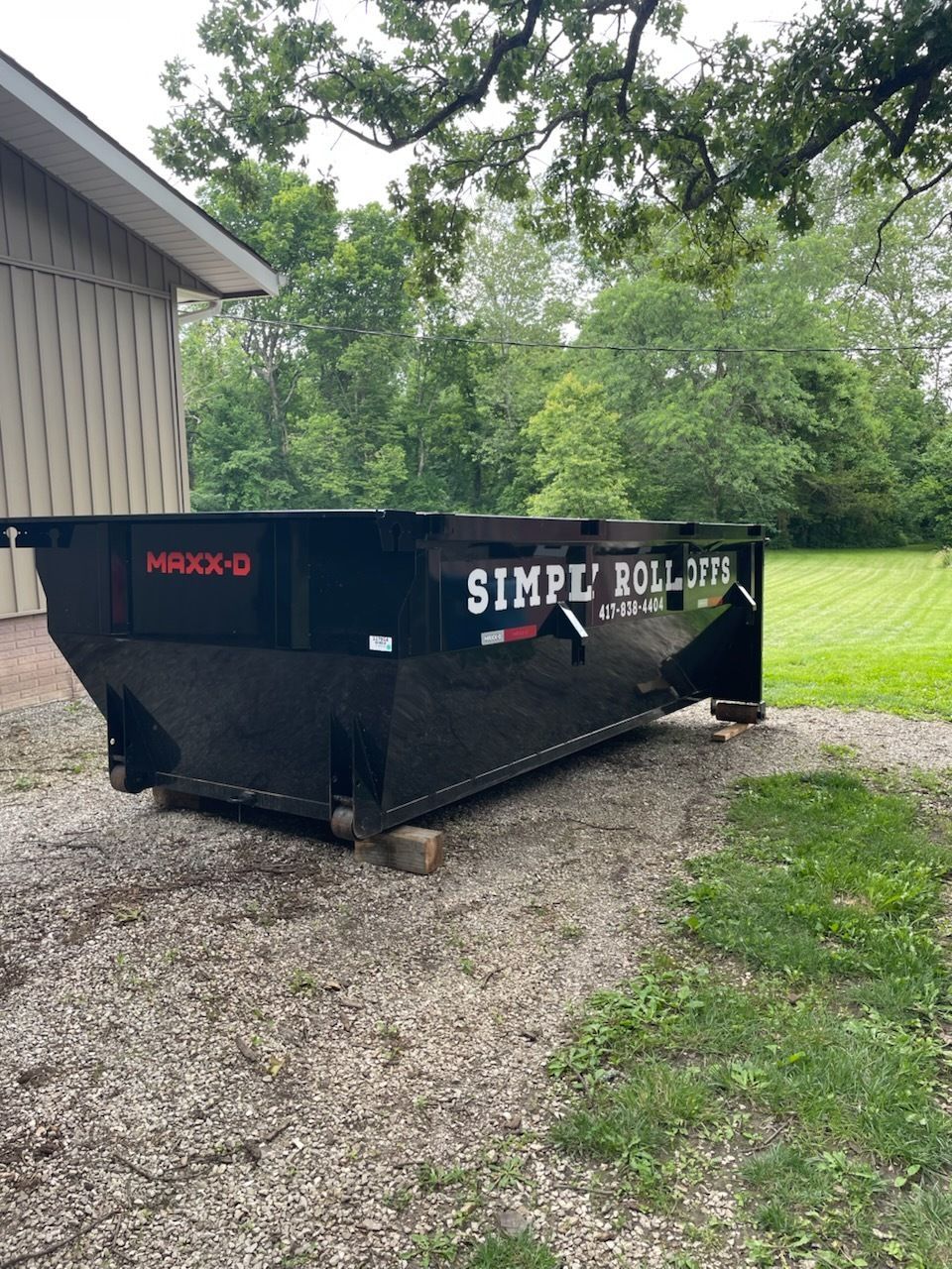 Black Simple Roll Off dumpster on gravel driveway near grass and trees.