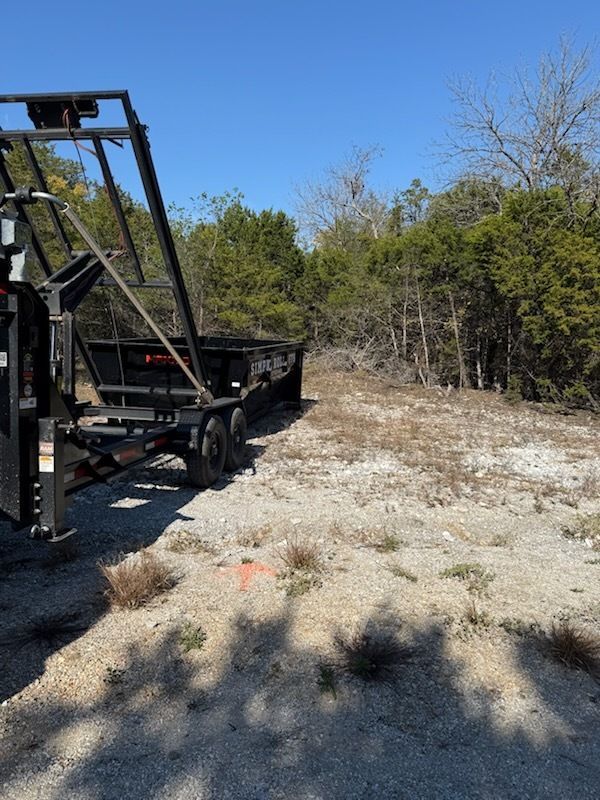 Black trailer parked on a gravel area with trees and a blue sky in the background.