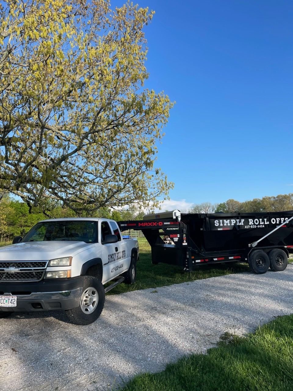 White truck with a black trailer on a gravel road, under a tree on a sunny day.