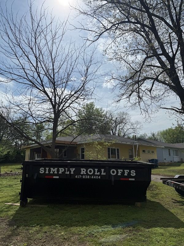 Black “Simply Roll Offs” dumpster in a grassy yard beneath leafless trees and a blue sky