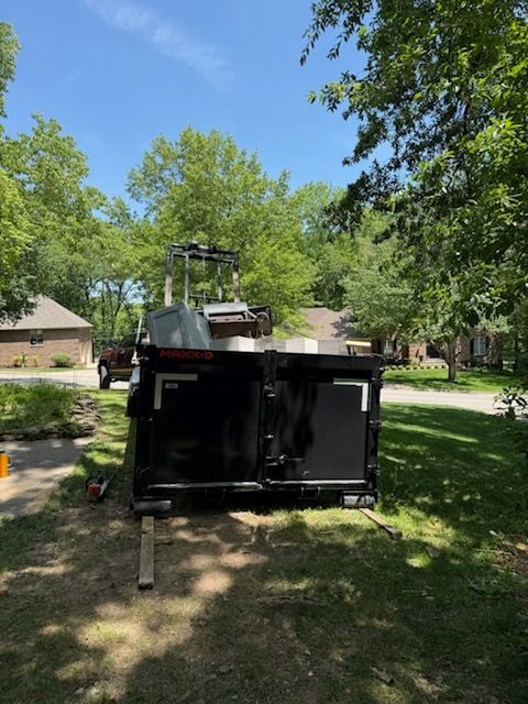Black dumpster in a grassy suburban yard with a small excavator behind it under trees and blue sky